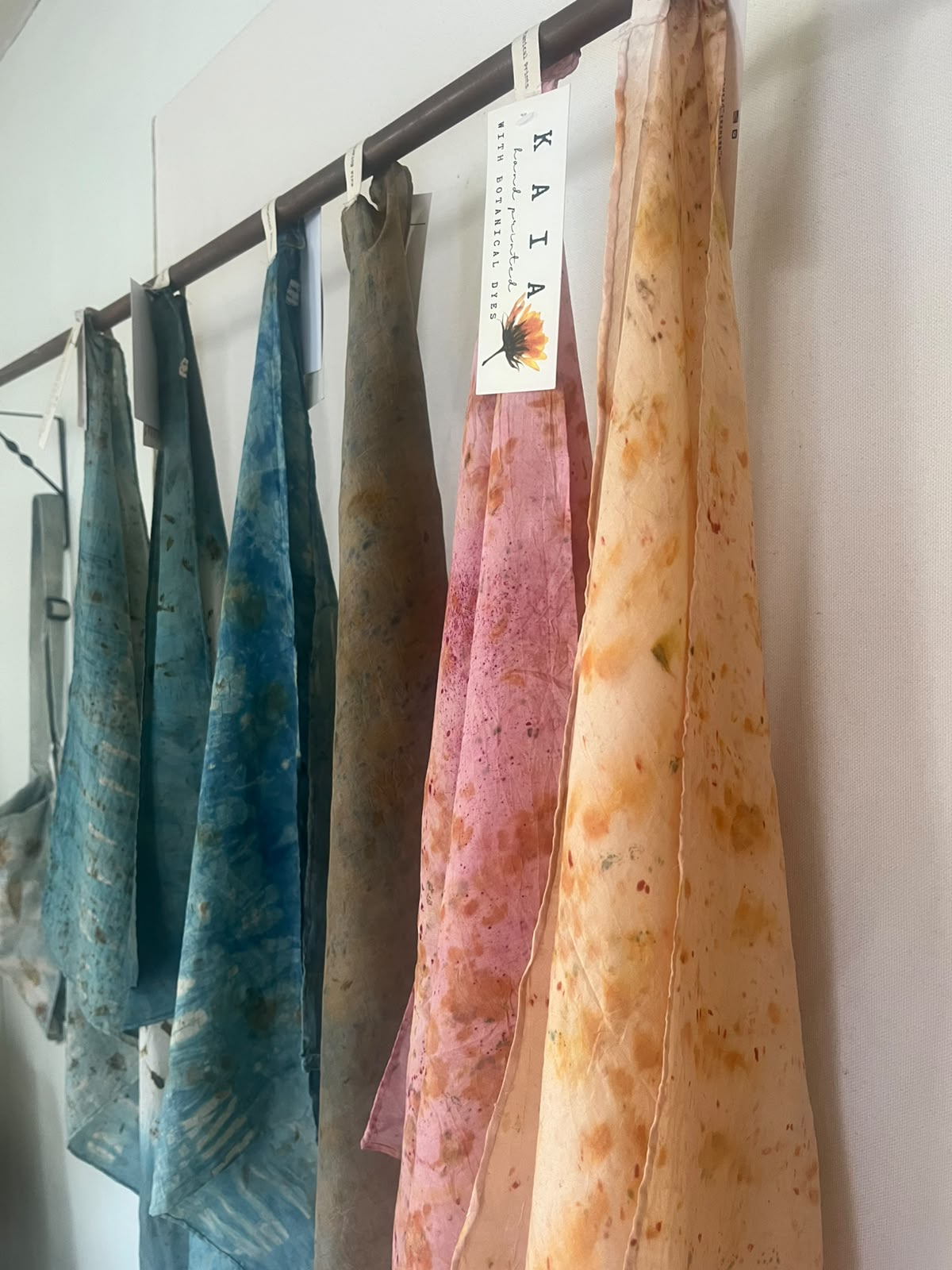Botanical dyed KAIA scarves in blue, olive, pink and orange hanging on a rail with a hand-written tag