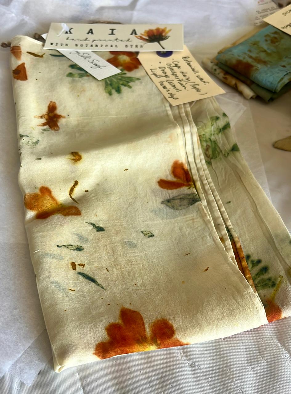A folded KAIA silk scarf printed with orange coreopsis and green leaves, with its hand-written botanical tag